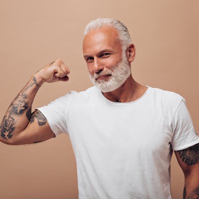 attractive-aged-man-in-white-outfit-demonstrates-muscles-on-beige-background-adult-with-gray-beard-in-tattoos-posing-into-camera