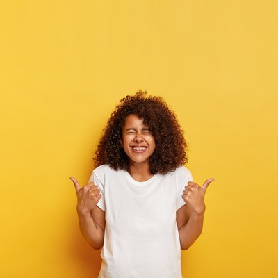 happy-overjoyed-ethnic-lady-with-bushy-crisp-hair-gives-positive-answer-with-thumbs-up-likes-awesome-idea-closes-eyes-from-laugh-dressed-mockup-t-shirt-isolated-yellow-wall