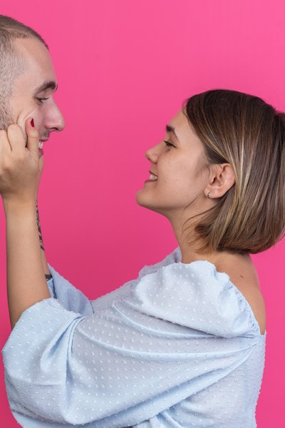 joyful-young-beautiful-couple-smiling-woman-pinching-cheeks-of-her-lovely-boyfriend-standing-over-pink-wall