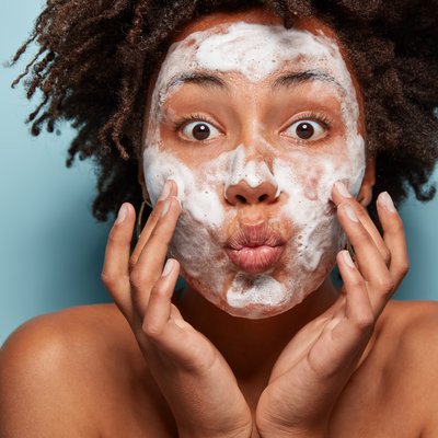 people-wellness-hygiene-skin-problems-concept-lovely-afro-american-lady-keeps-lips-folded-touches-cheeks-has-white-foam-faces-washes-with-beauty-gel-feels-refreshed-has-wide-opened-eyes