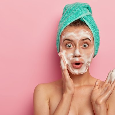 photo-surprised-european-woman-washes-face-with-foam-gel-wants-have-refreshed-well-cared-skin-stands-topless-wears-wrapped-towel-wet-hair-poses-against-pink-background-free-space-aside