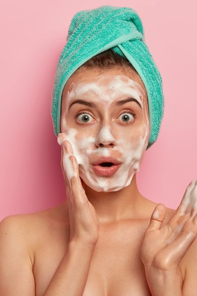 photo-surprised-european-woman-washes-face-with-foam-gel-wants-have-refreshed-well-cared-skin-stands-topless-wears-wrapped-towel-wet-hair-poses-against-pink-background-free-space-aside